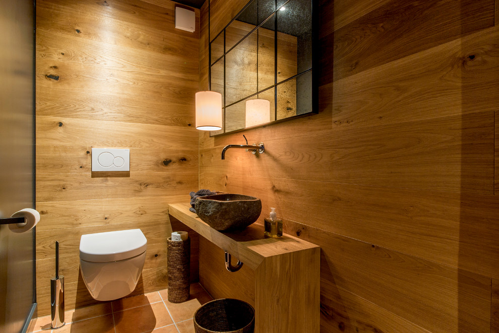 Photo of a small rustic cloakroom in Hanover with a wall mounted toilet, a vessel sink, wooden worktops, medium wood cabinets, black walls, terracotta flooring and red floors.