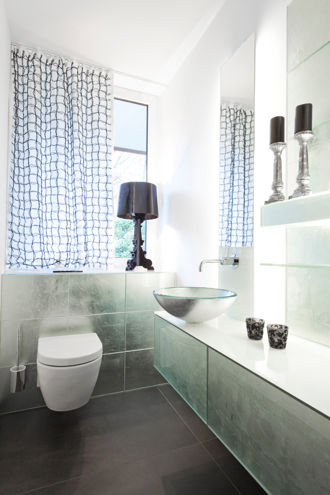 Exemple d'un petit WC suspendu éclectique avec une vasque, un placard à porte plane, un mur blanc, des carreaux de miroir, un plan de toilette en verre et un carrelage gris.