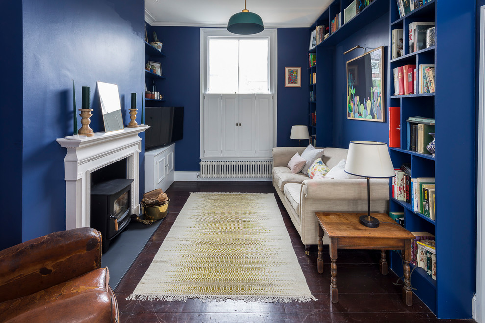 Modelo de sala de estar con biblioteca abierta clásica renovada grande con paredes azules, suelo de madera oscura, estufa de leña, marco de chimenea de yeso, televisor colgado en la pared y suelo marrón