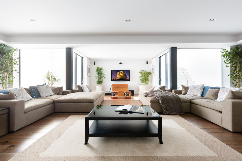 This is an example of a contemporary open plan games room in London with white walls, medium hardwood flooring, a wall mounted tv and brown floors.