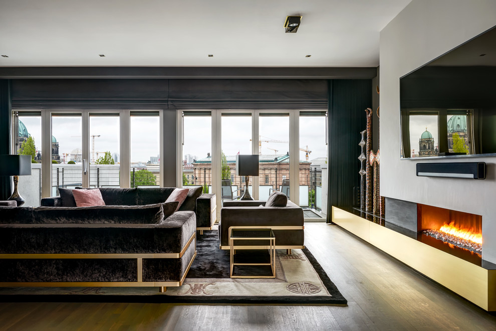 Diseño de sala de estar tipo loft contemporánea grande con paredes grises, suelo de madera en tonos medios, chimenea lineal, marco de chimenea de yeso, televisor colgado en la pared y suelo marrón