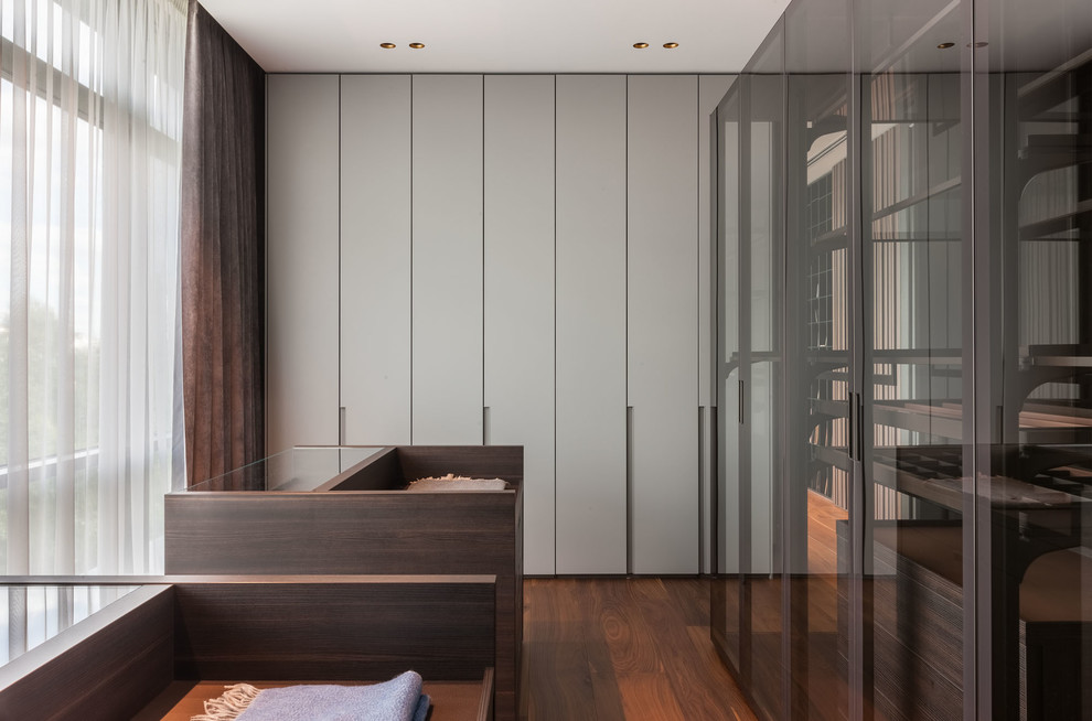 Réalisation d'un dressing room design neutre avec un placard à porte plane, des portes de placard grises, un sol en bois brun et un sol marron.