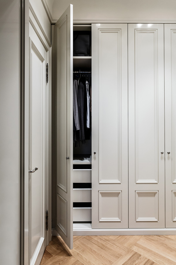 На фото: гардеробная унисекс в классическом стиле с фасадами с утопленной филенкой, белыми фасадами и светлым паркетным полом с