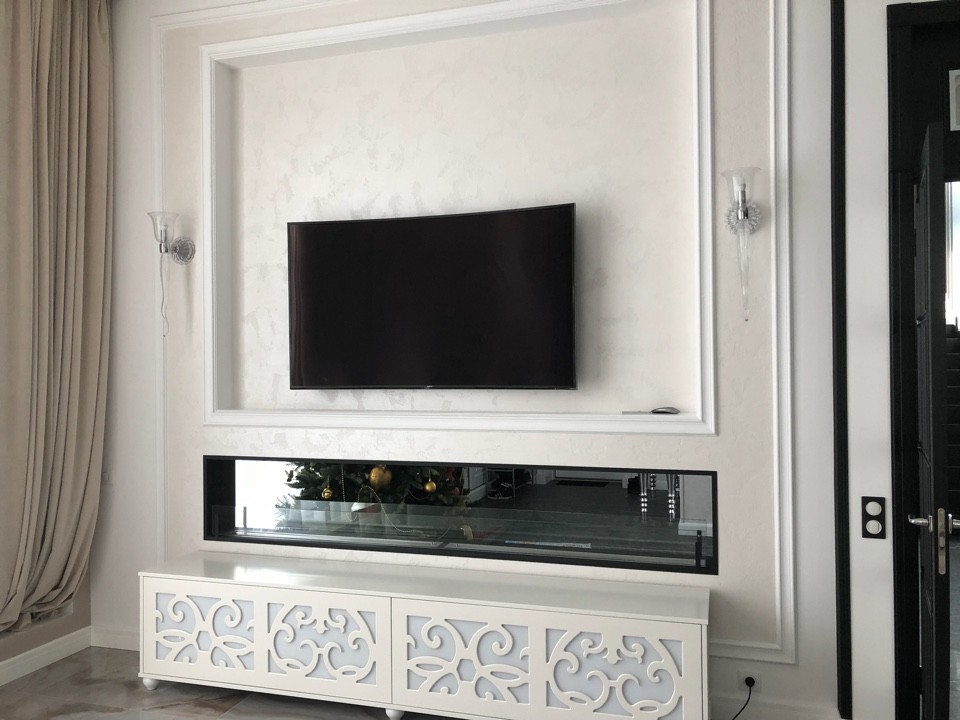 Modelo de salón grande con paredes blancas, chimenea de doble cara, televisor colgado en la pared y suelo beige
