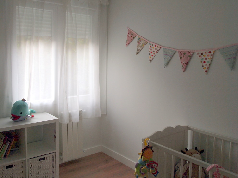 Diseño de habitación de bebé niña escandinava de tamaño medio con paredes blancas, suelo laminado y suelo marrón