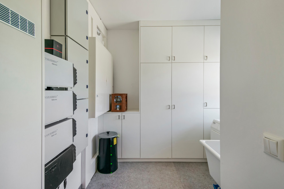 This is an example of a medium sized contemporary l-shaped utility room in Hamburg with a single-bowl sink, flat-panel cabinets, white cabinets, white walls, vinyl flooring, a side by side washer and dryer, grey floors and white worktops.
