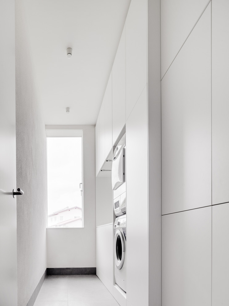 Design ideas for a medium sized modern single-wall utility room in Berlin with flat-panel cabinets, white cabinets, white walls and a stacked washer and dryer.