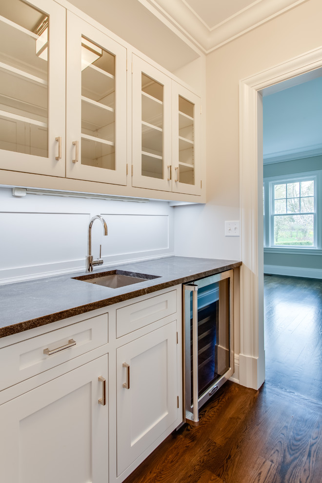 Inredning av en klassisk stor u-formad hemmabar med vask, med en undermonterad diskho, luckor med glaspanel, vita skåp, marmorbänkskiva, vitt stänkskydd, mellanmörkt trägolv och brunt golv