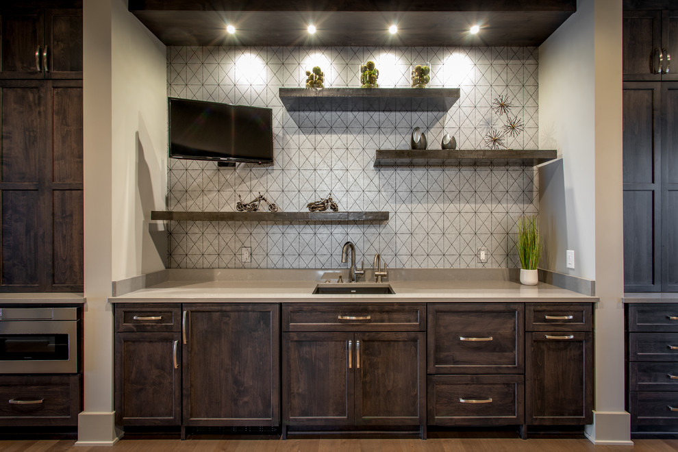 Klassisk inredning av en mellanstor grå linjär grått hemmabar med vask, med en undermonterad diskho, luckor med infälld panel, skåp i mörkt trä, bänkskiva i kvarts, grått stänkskydd, stänkskydd i keramik, mellanmörkt trägolv och brunt golv