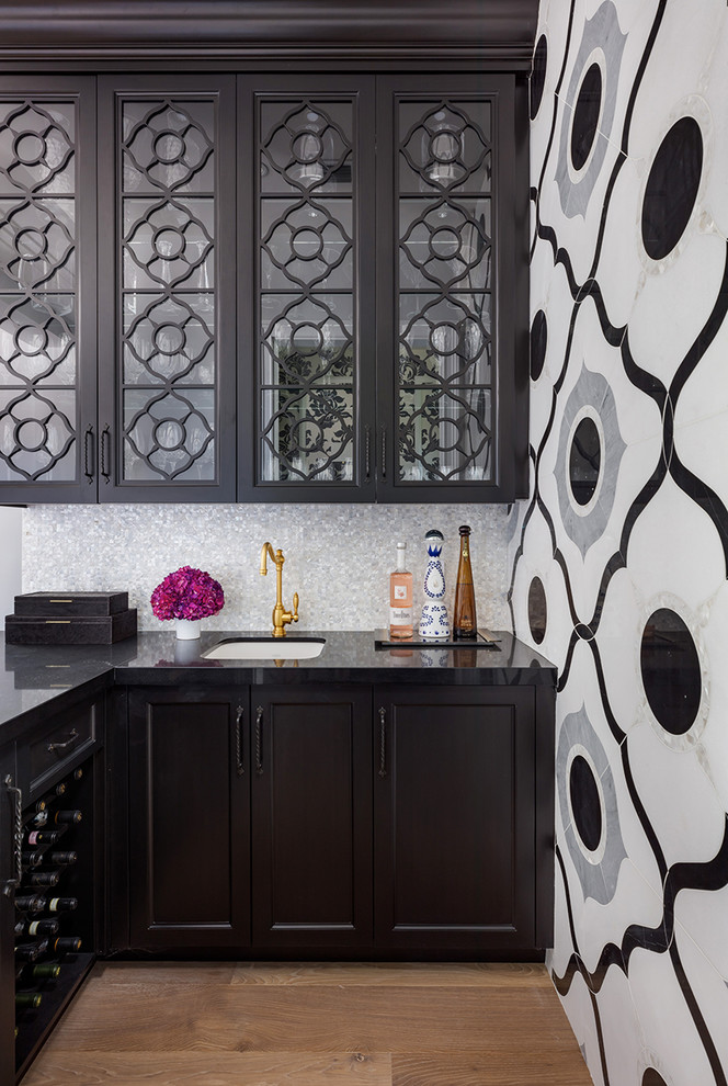 Design ideas for a classic l-shaped wet bar in Other with a submerged sink, glass-front cabinets, black cabinets, light hardwood flooring, black worktops and a feature wall.