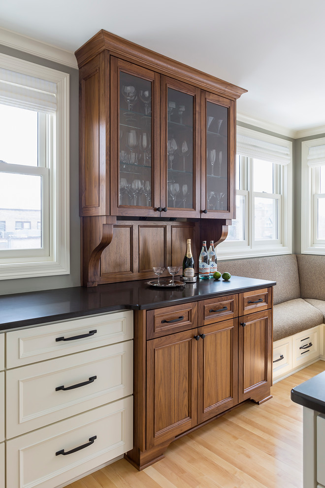 This is an example of a traditional l-shaped home bar in Minneapolis with a submerged sink, recessed-panel cabinets, medium wood cabinets, engineered stone countertops, white splashback and light hardwood flooring.