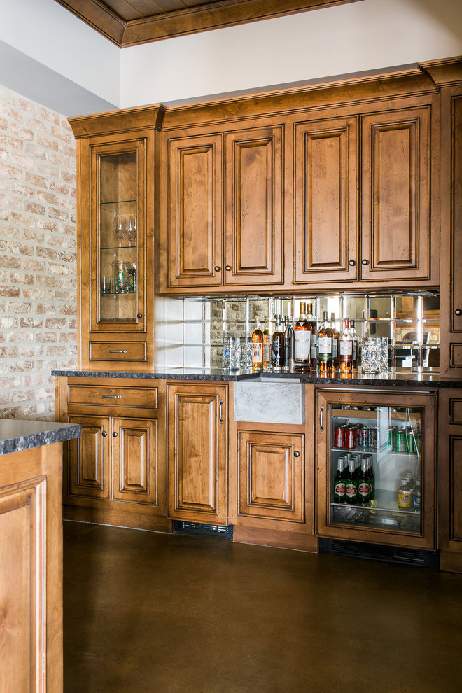 Immagine di un angolo bar con lavandino tradizionale di medie dimensioni con ante con bugna sagomata, ante in legno scuro, top in vetro, paraspruzzi a specchio e pavimento in cemento