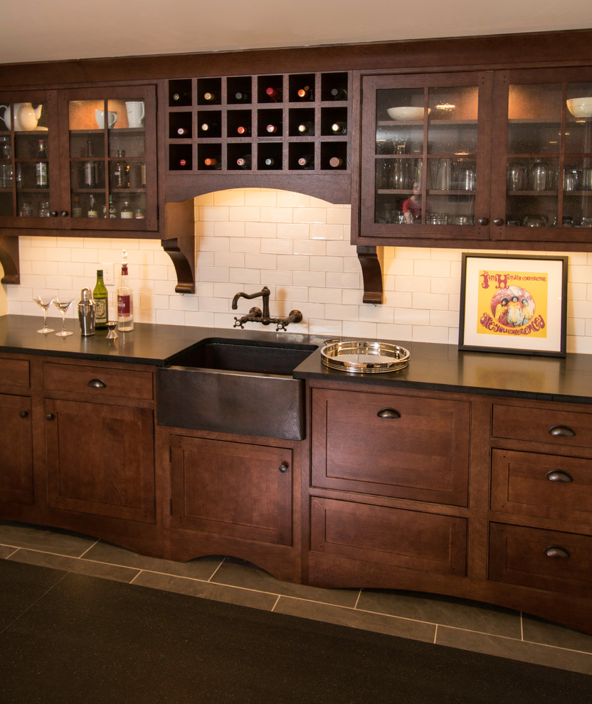 This is an example of a small traditional galley wet bar in New York with a built-in sink, beaded cabinets, brown cabinets, soapstone worktops, white splashback, ceramic splashback, medium hardwood flooring, brown floors and black worktops.