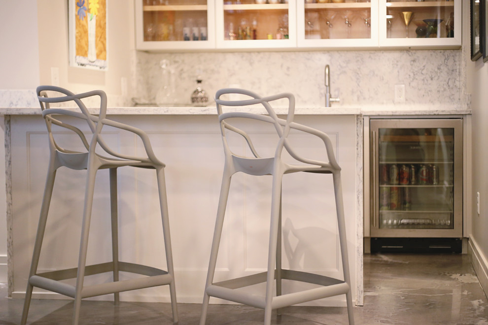 Modern inredning av en mellanstor parallell hemmabar med stolar, med en undermonterad diskho, luckor med glaspanel, vita skåp, marmorbänkskiva, vitt stänkskydd, stänkskydd i sten och betonggolv