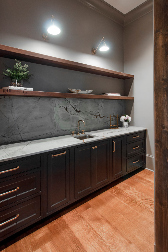Inredning av en mellanstor grå parallell grått hemmabar med vask, med en undermonterad diskho, luckor med profilerade fronter, svarta skåp, bänkskiva i kvartsit, grått stänkskydd, mörkt trägolv och brunt golv