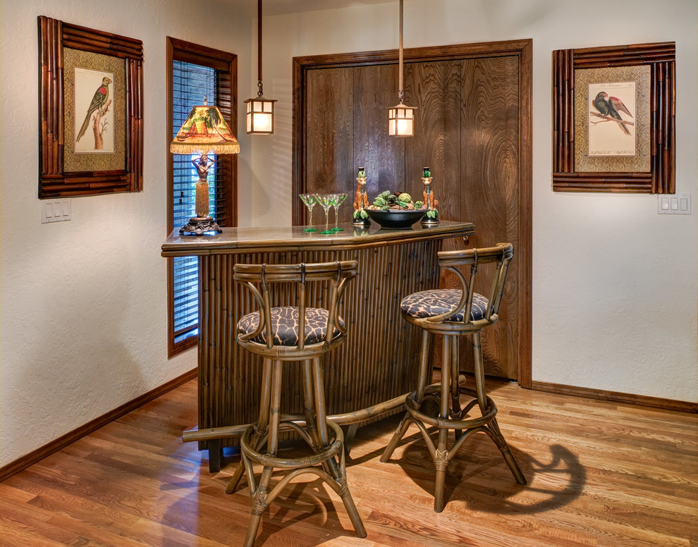 Aménagement d'un bar de salon exotique de taille moyenne avec des tabourets, des portes de placard marrons, un plan de travail en verre et un sol en bois brun.