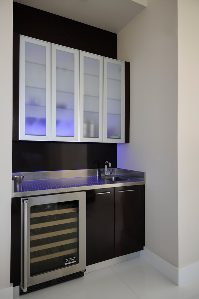 Inspiration for a small contemporary single-wall wet bar in Miami with an integrated sink, glass-front cabinets, brown cabinets, stainless steel worktops, brown splashback, glass sheet splashback, ceramic flooring and white floors.