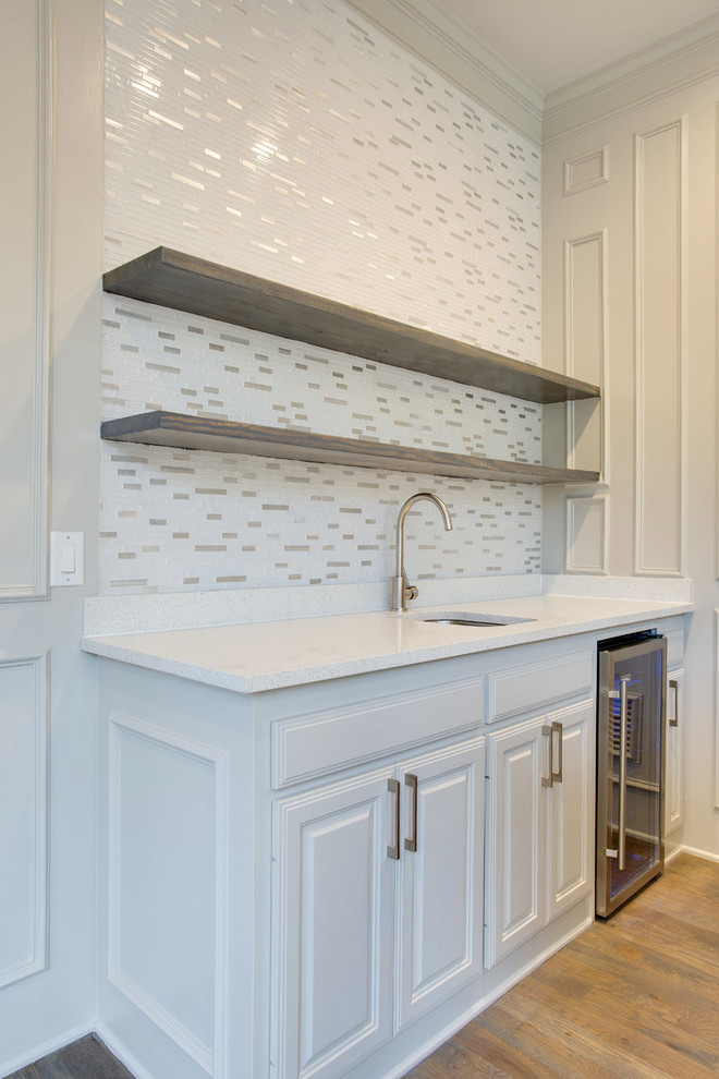 Design ideas for a large traditional galley wet bar in Dallas with a submerged sink, raised-panel cabinets, white cabinets, engineered stone countertops, white splashback, mosaic tiled splashback, medium hardwood flooring, grey floors and white worktops.