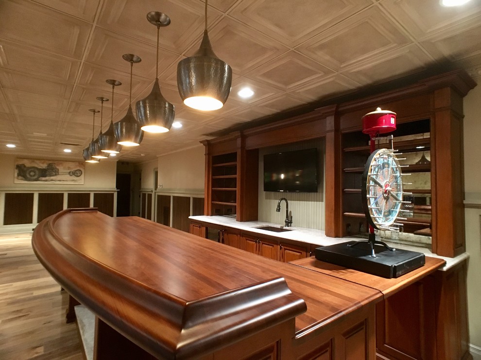 Diseño de bar en casa con barra de bar lineal, tradicional, extra grande, con armarios con paneles con relieve, puertas de armario de madera oscura, encimera de madera, suelo de madera clara y fregadero bajoencimera