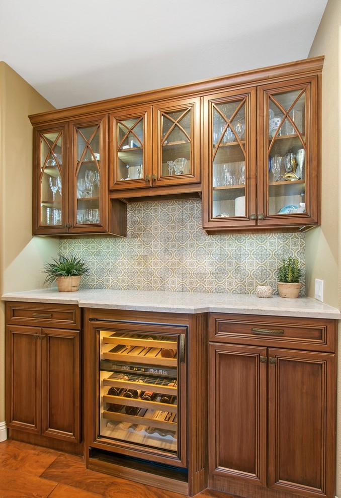 Inspiration for a small transitional galley medium tone wood floor and brown floor home bar remodel in San Diego with no sink, recessed-panel cabinets, medium tone wood cabinets, quartz countertops, gray backsplash and travertine backsplash