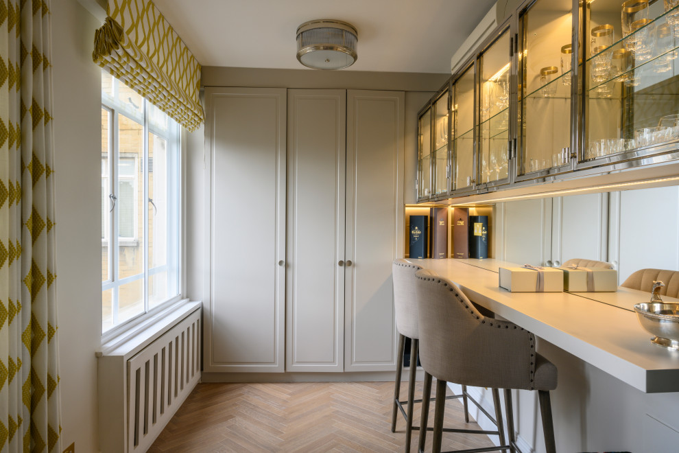 Photo of a medium sized contemporary l-shaped breakfast bar in London with raised-panel cabinets, grey cabinets, mirror splashback, medium hardwood flooring, beige floors and grey worktops.