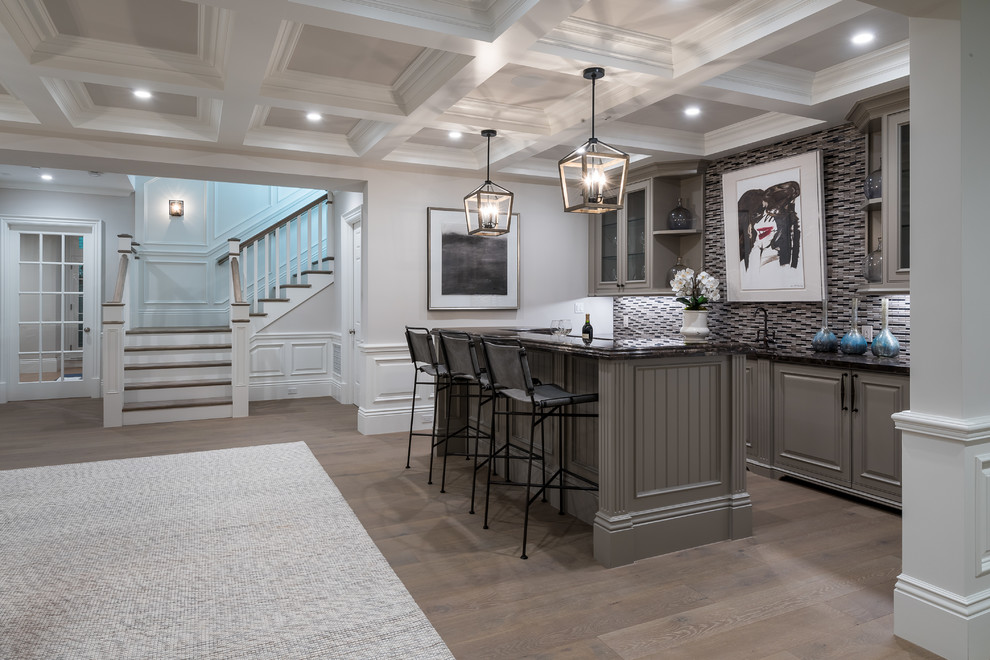 Transitional galley medium tone wood floor and brown floor home bar photo in Los Angeles with raised-panel cabinets, gray cabinets, gray backsplash, mosaic tile backsplash and black countertops