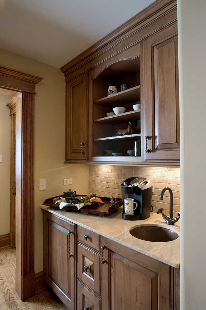 This is an example of a small traditional galley wet bar in Milwaukee with a submerged sink, raised-panel cabinets, medium wood cabinets, limestone worktops, beige splashback, medium hardwood flooring and travertine splashback.