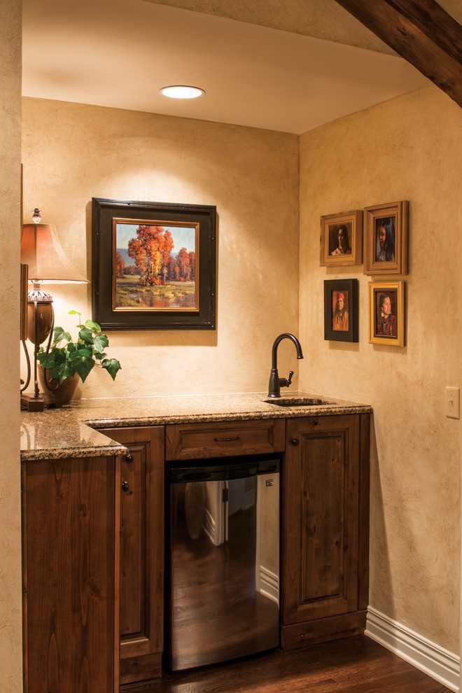 Klassische Hausbar mit Unterbauwaschbecken, profilierten Schrankfronten, Granit-Arbeitsplatte, braunem Holzboden und Bartresen in Wichita