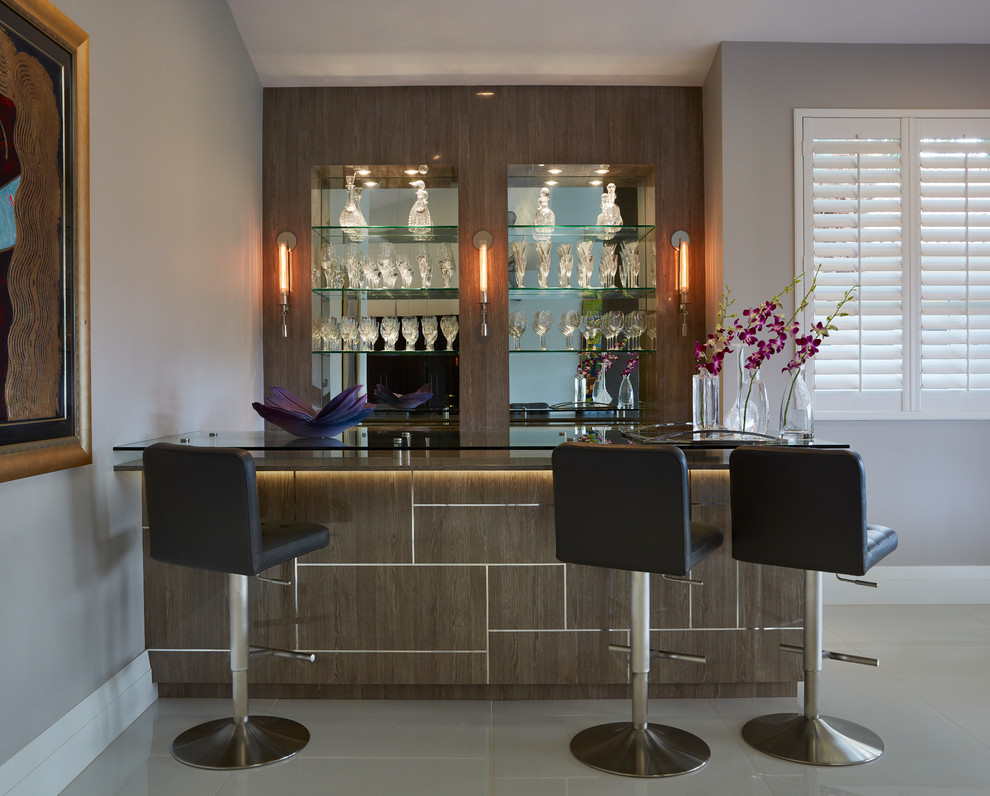 Réalisation d'un bar de salon parallèle design en bois brun de taille moyenne avec des tabourets, un plan de travail en verre, une crédence miroir, un placard à porte plane et un sol blanc.