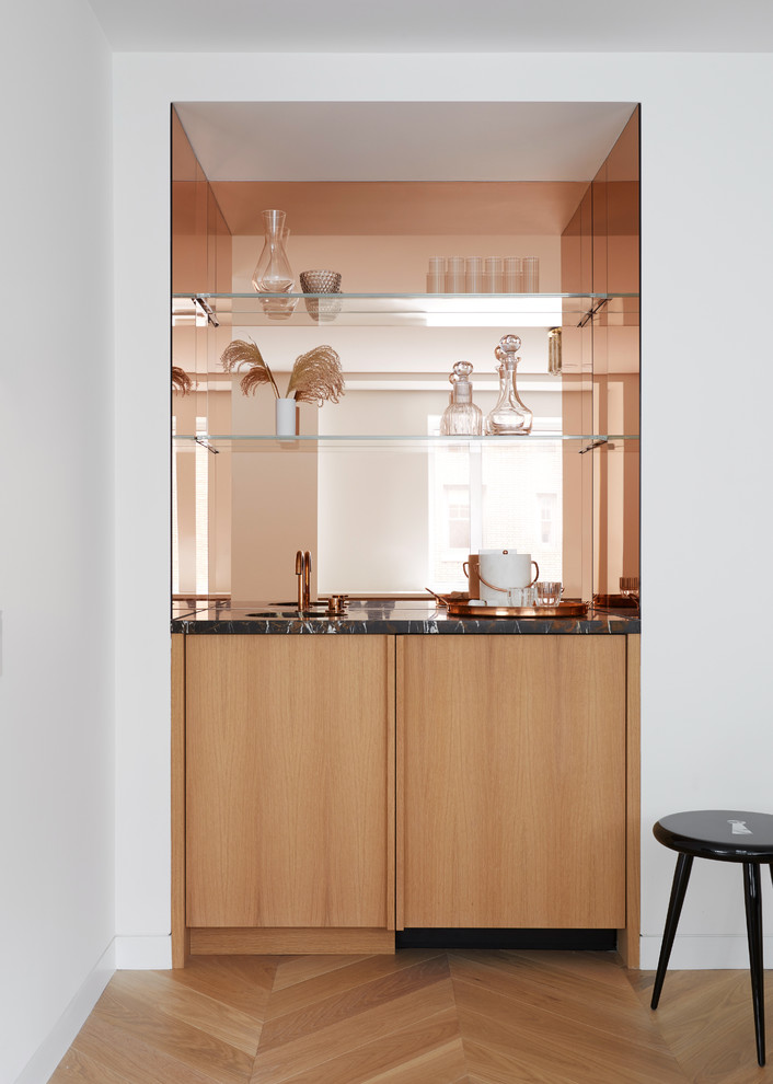 Contemporary single-wall wet bar in New York with flat-panel cabinets, light wood cabinets, mirror splashback, light hardwood flooring, beige floors and black worktops.