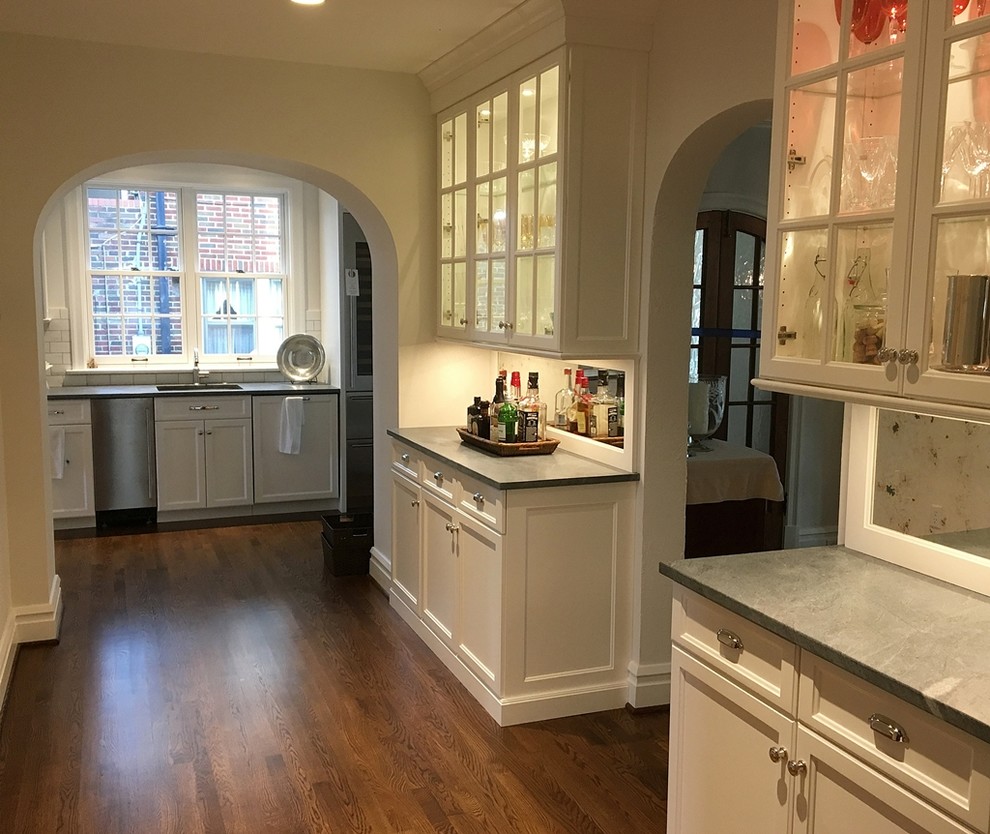 Klassisk inredning av en stor linjär hemmabar med vask, med vita skåp, mellanmörkt trägolv, luckor med glaspanel, bänkskiva i täljsten och spegel som stänkskydd