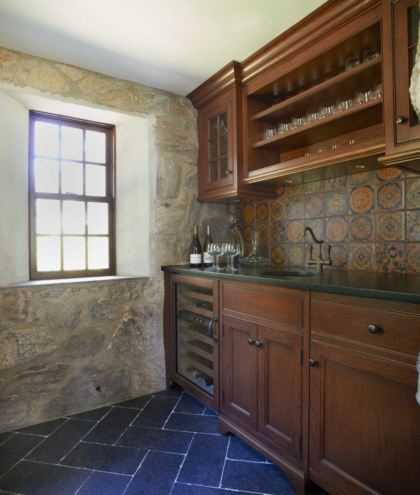 Inredning av en klassisk linjär hemmabar med vask, med en undermonterad diskho, släta luckor, skåp i mellenmörkt trä, granitbänkskiva, flerfärgad stänkskydd, stänkskydd i keramik och kalkstensgolv