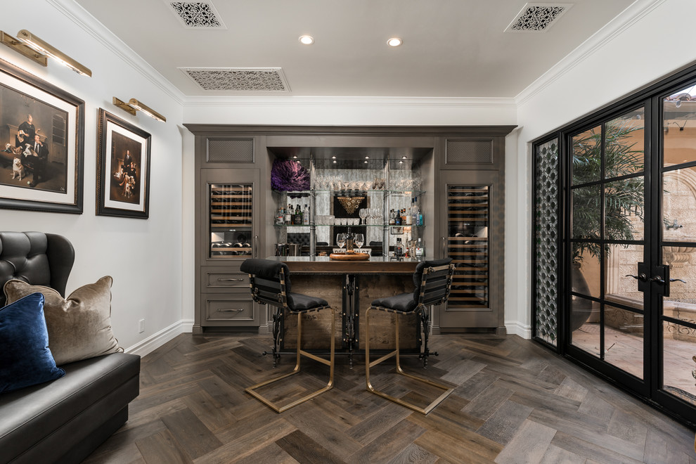 Cette photo montre un bar de salon parallèle méditerranéen avec des tabourets, un placard avec porte à panneau encastré, des portes de placard grises, une crédence miroir, parquet foncé et un sol marron.