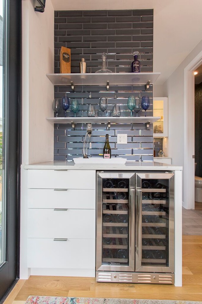 Inspiration for a small contemporary single-wall home bar in Denver with flat-panel cabinets, white cabinets, quartz worktops, blue splashback, glass tiled splashback, light hardwood flooring and brown floors.