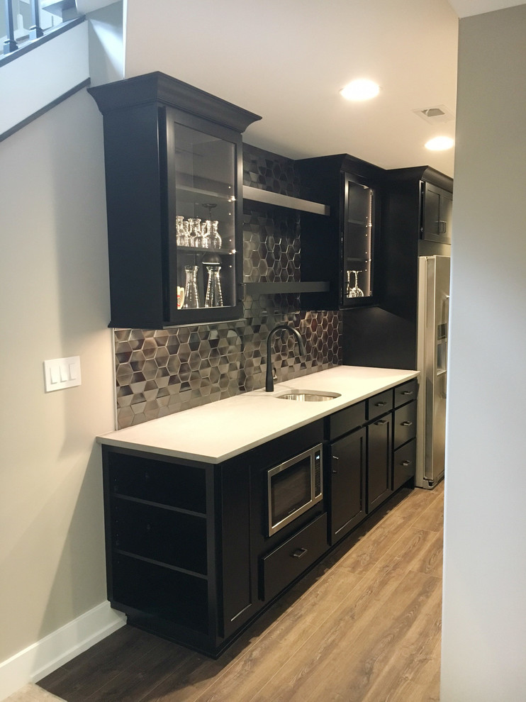 This is an example of a large modern galley wet bar in Other with a submerged sink, shaker cabinets, black cabinets, engineered stone countertops, grey splashback, metal splashback, light hardwood flooring and grey floors.