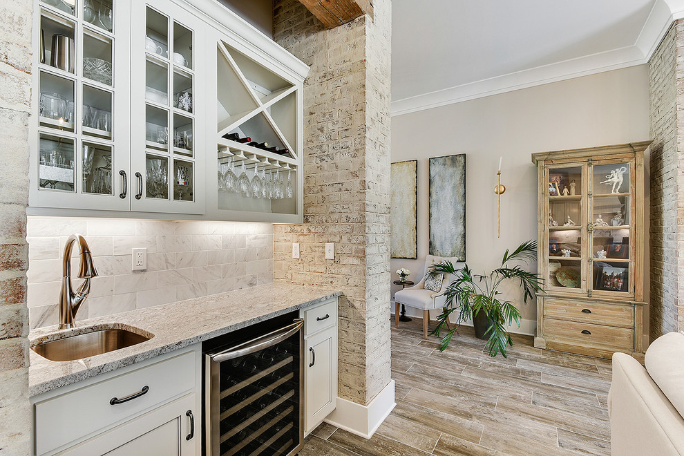 Inredning av en klassisk liten linjär hemmabar med vask, med en undermonterad diskho, luckor med glaspanel, vita skåp, granitbänkskiva, vitt stänkskydd, stänkskydd i marmor, klinkergolv i porslin och brunt golv