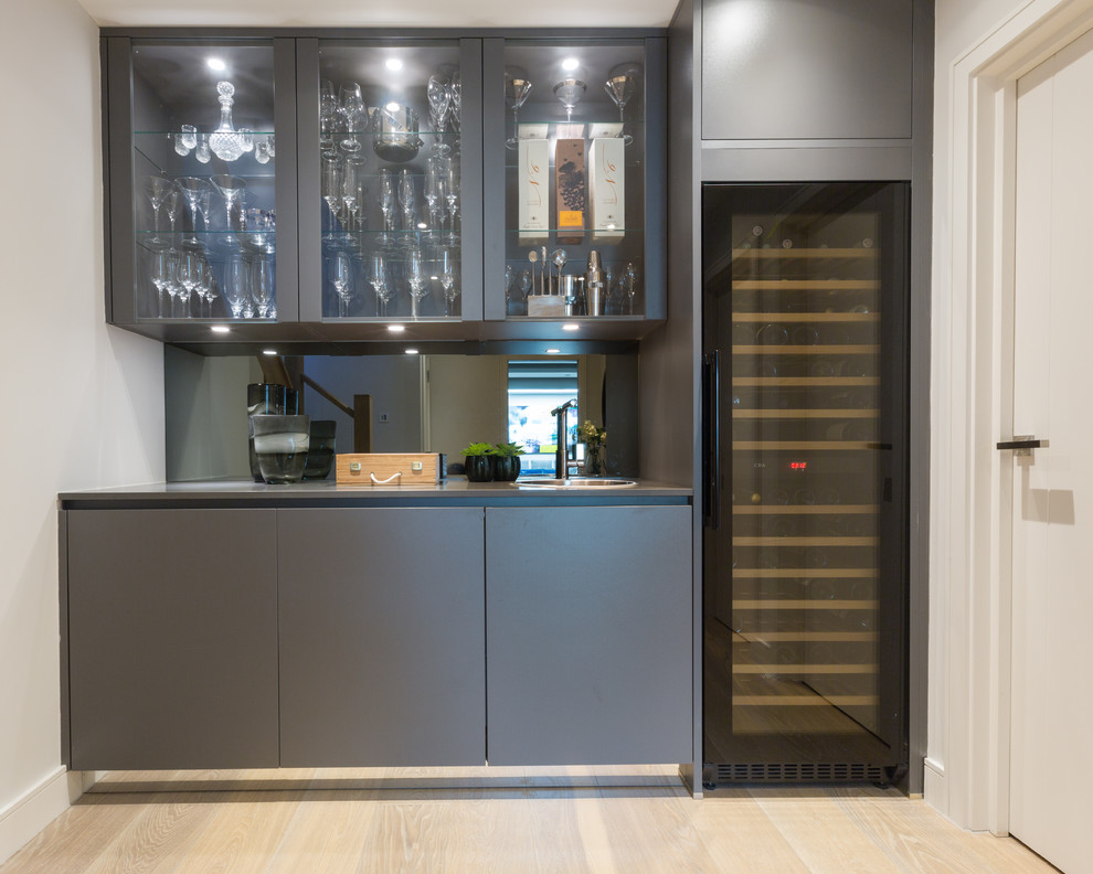 Small contemporary single-wall wet bar in Gloucestershire with a built-in sink, flat-panel cabinets, grey cabinets, composite countertops, grey splashback, mirror splashback, light hardwood flooring and grey worktops.