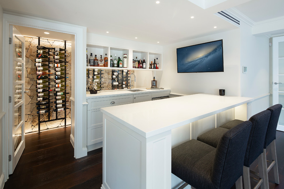 Cette photo montre un bar de salon bord de mer de taille moyenne avec des tabourets, un évier encastré, des portes de placard blanches, un plan de travail en quartz modifié, parquet foncé, un sol marron, un plan de travail blanc, un placard sans porte et une crédence en feuille de verre.