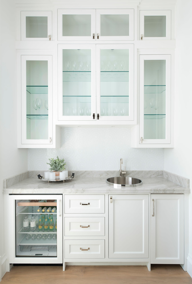 Mediterranean single-wall wet bar in Dallas with light hardwood flooring, a built-in sink, glass-front cabinets, white cabinets, beige floors and grey worktops.