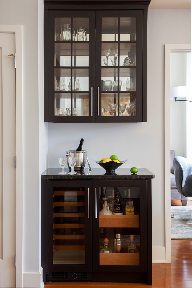 Idee per un piccolo angolo bar design con ante di vetro, ante in legno bruno, top in granito e parquet chiaro