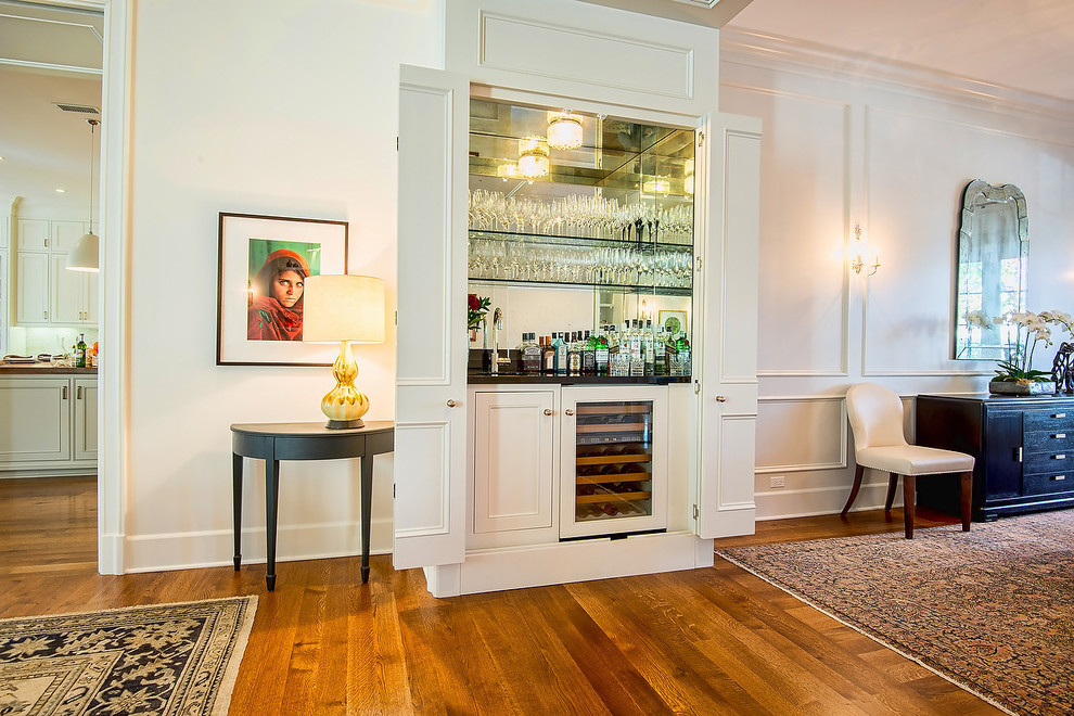 Inspiration pour un petit bar de salon avec évier linéaire traditionnel avec un sol en bois brun, un placard avec porte à panneau encastré, des portes de placard blanches et une crédence miroir.