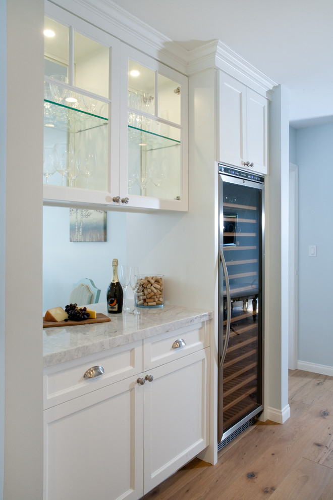 This is an example of a large beach style home bar in Los Angeles with shaker cabinets, white cabinets, marble worktops, blue splashback, medium hardwood flooring and beige floors.