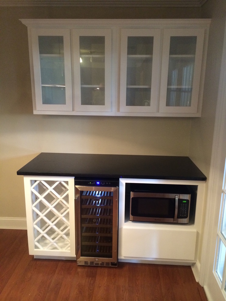 This is an example of a small classic single-wall wet bar in Other with medium hardwood flooring, brown floors, no sink, glass-front cabinets, white cabinets and composite countertops.