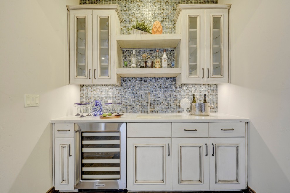 This is an example of a medium sized traditional single-wall wet bar in Austin with a submerged sink, raised-panel cabinets, distressed cabinets, engineered stone countertops, blue splashback, glass tiled splashback, porcelain flooring and brown floors.
