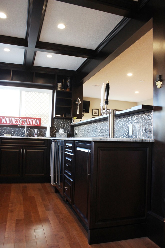 Example of a mid-sized classic u-shaped medium tone wood floor seated home bar design in Calgary with an undermount sink, raised-panel cabinets, dark wood cabinets, granite countertops, gray backsplash and glass tile backsplash