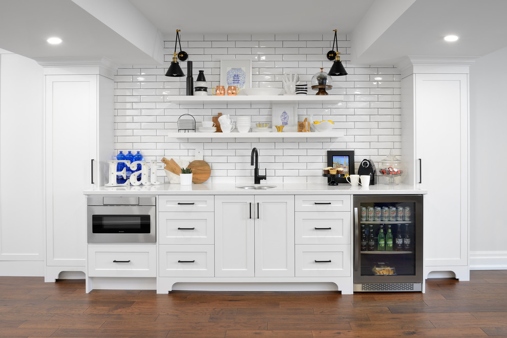 Cette image montre un bar de salon avec évier linéaire traditionnel de taille moyenne avec un évier encastré, un placard à porte shaker, des portes de placard blanches, un plan de travail en quartz, une crédence blanche, une crédence en carrelage métro, parquet foncé et un plan de travail blanc.