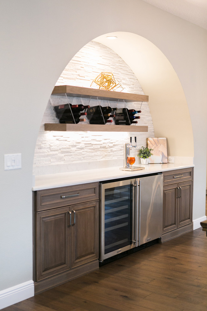 グランドラピッズにあるトランジショナルスタイルのおしゃれなホームバー (I型、レイズドパネル扉のキャビネット、中間色木目調キャビネット、白いキッチンパネル、無垢フローリング、茶色い床、白いキッチンカウンター) の写真