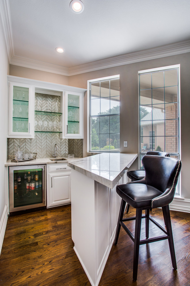 Traditional single-wall wet bar in Dallas with a submerged sink, glass-front cabinets, white cabinets, beige splashback, medium hardwood flooring, brown floors and beige worktops.