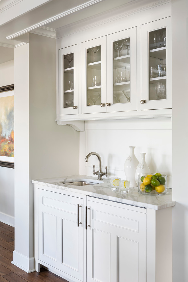 Klassisk inredning av en mellanstor vita vitt hemmabar, med en undermonterad diskho, luckor med infälld panel, vita skåp, marmorbänkskiva, grått stänkskydd, mörkt trägolv och stänkskydd i trä