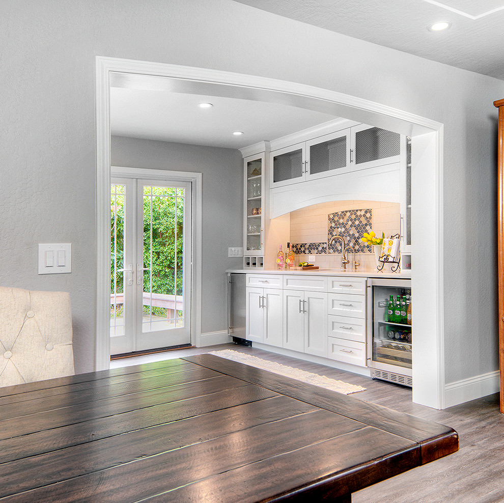 This is an example of a medium sized traditional single-wall wet bar in San Francisco with a submerged sink, shaker cabinets, white cabinets, engineered stone countertops, multi-coloured splashback, mosaic tiled splashback, vinyl flooring, brown floors and white worktops.